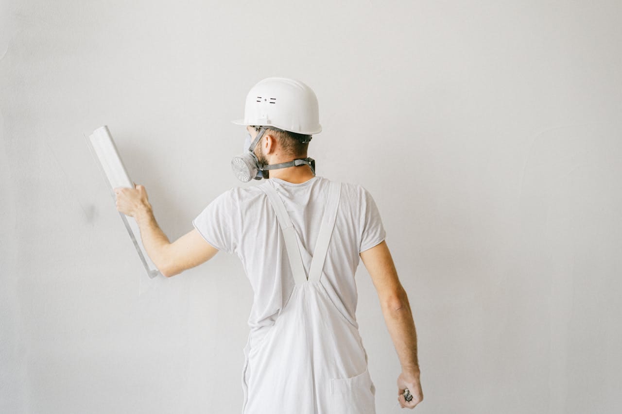 services-03 Back view of a construction worker in safety gear plastering a white wall.