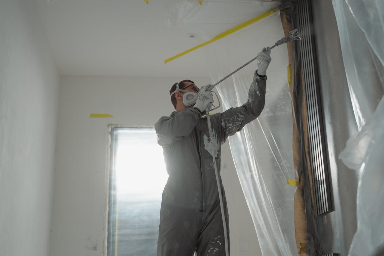 about-01 A professional painter wearing protective gear spray paints a wall indoors, working on a home renovation project.