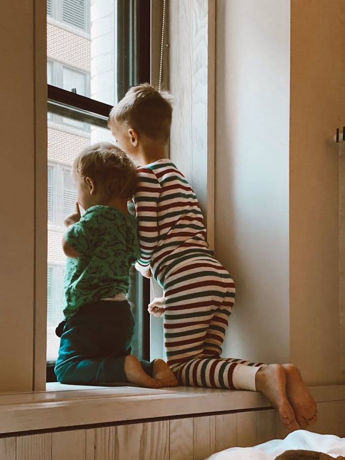 missions-03 Two kids in pajamas curiously peering out of a window inside their home.