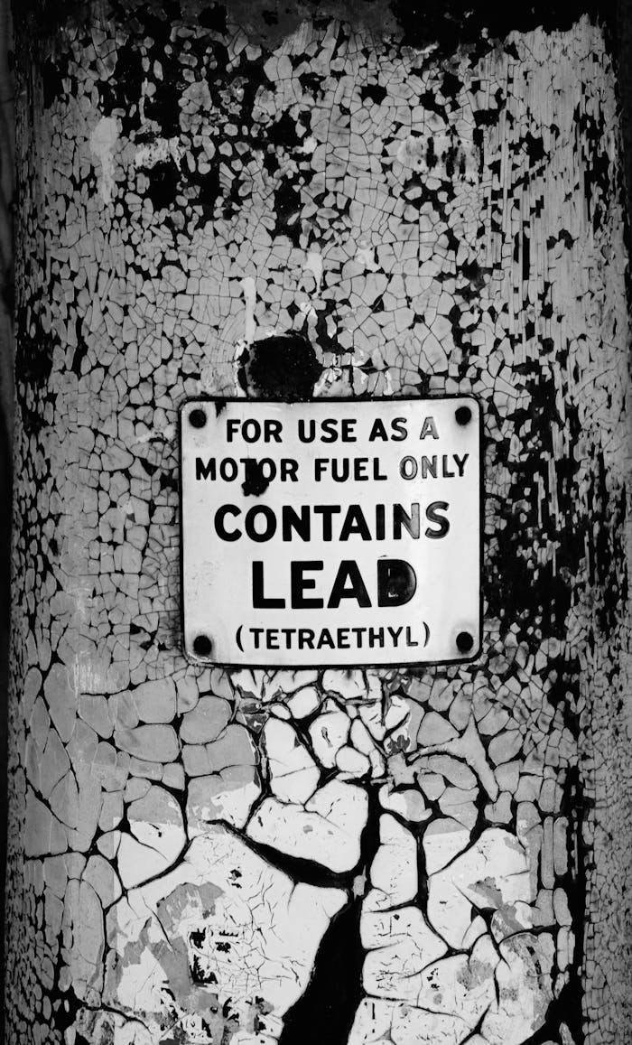 hero-02 Close-up of a vintage warning sign indicating lead content for motor fuel, mounted on a weathered wall.